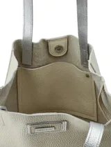Cartera tipo tote de cuero color crudo con base y asas plateadas, de diseño vertical y rectangular con detalle de placa metálica con logo de la marca en el frente.