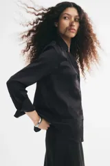 Blusa negra de tela brillante, con cuello clásico, escote en V con botones frontales, mangas largas con puños sencillos y bajo redondeado.