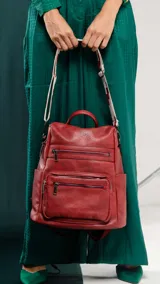 Mochila de símil cuero color marrón con apertura superior y tres bolsillos frontales con cierres metálicos. Cuenta con bolsillos laterales y una correa de galón con diseño geométrico, regulable y extraíble, que permite su uso como bandolera.