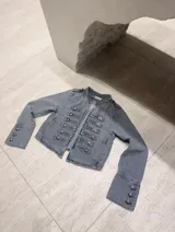 Chaqueta de jean color gris estilo militar con diseño napoleónico, cierre frontal de ganchos y múltiples botones metálicos ornamentales en la parte delantera y en los puños.