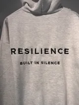 Canguro gris melange con capucha, confeccionado en algodón con felpa. Presenta bordado en color negro en la espalda con la frase 'RESILIENCE BUILT IN SILENCE'.