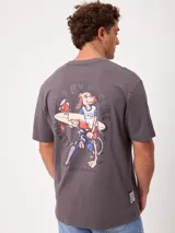 Remera gris de corte loose fit, manga corta y cuello redondo. Cuenta con un gran estampado en la espalda de un perro en bicicleta con una tabla de surf, acompañado de un loro y la leyenda 'Rusty Worldwide'.