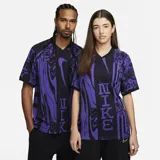 Remera deportiva Nike de la línea Culture of Football, con diseño estampado en tonos negros y violetas inspirado en el fútbol, cuello en V y mangas cortas.
