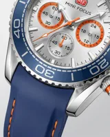 Reloj de pulsera analógico con cronógrafo, caja de metal y correa de caucho azul. Presenta esfera blanca con detalles en naranja y bisel giratorio azul.