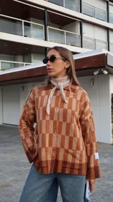 Buzo tipo saco abotonado con diseño de cuadros en tonos marrones y beige, corte recto y oversize. Presenta cuello camisero, cierre frontal con botones personalizados y dos bolsillos delanteros.