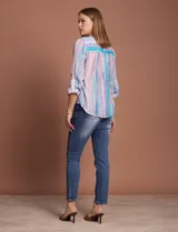 Blusa de gasa con estampado de rayas verticales en tonos celestes, violetas y blancos. Presenta cuello mao con escote en V, cierre frontal con botones, bolsillo aplicado en el pecho y mangas largas con presilla para regular el largo. El diseño incluye un nudo en la parte inferior delantera.