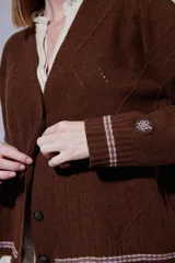 Saco tejido en lambswool con diseño de rombos y trenzas en el frente. Presenta cierre frontal con botones, cuello en V y detalles de rayas en contratono en puños y ruedo. Incluye pin bordado en la manga.