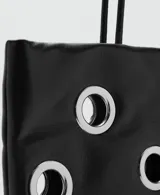Bolso tipo shopper de color negro con diseño de ojales metálicos plateados. Cuenta con doble asa de hombro y cierre de botón magnético.