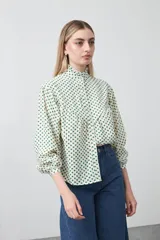 Camisa de viscosa con estampado de lunares verdes sobre fondo crema, cuello alto con volados y cierre frontal de botones. Presenta mangas largas abullonadas con puños elásticos y un corte holgado con frunces que aportan volumen.