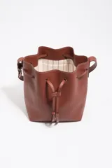 Bolso tipo saco de cuero marrón con cierre de cordón ajustable, asa larga regulable y logotipo de BIMBA Y LOLA impreso en el frente con letras blancas.