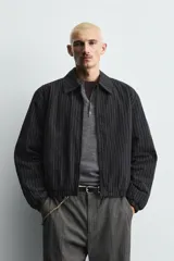 Cazadora de hombre de ajuste cropped fit, confeccionada en tejido de algodón con patrón de rayas verticales finas en tonos oscuros (negro/azul marino y marrón/beige). Presenta cuello solapa, cierre frontal de cremallera, bolsillos laterales y elástico en puños y bajo.