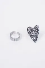 Set de dos anillos de metal plateado. Uno presenta un diseño de banda texturizada con pequeños eslabones, mientras que el otro destaca por un dije central en forma de corazón con una superficie irregular y brillante.