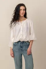 Blusa blanca de diseño amplio, con mangas abullonadas, escote redondo fruncido y cierre frontal de botones nacarados. Cuenta con elástico en la cintura que genera efecto blusón.