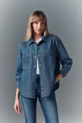 Camisa de denim azul con apliques metálicos plateados y bordado de piedras rojas y negras en los bolsillos delanteros. Tiene calce amplio, cuello camisero y cierre con botones a presión.