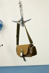 Bandolera de tela color verde militar, con tapa frontal y hebilla metálica. Cuenta con correa regulable y bolsillos laterales con cierre.