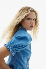 Blusa corta confeccionada en denim de algodón, con diseño entallado, cuello con volados, mangas cortas abullonadas con puños elásticos y botonadura frontal. Presenta alforzas decorativas en el frente y un volado en el ruedo.