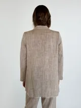 Blazer de lino y algodón en tono beige con textura a rayas verticales sutiles. Presenta solapas clásicas, un solo botón frontal marrón y un corte ligeramente holgado. Los bordes de las mangas y el ruedo tienen un acabado deshilachado.
