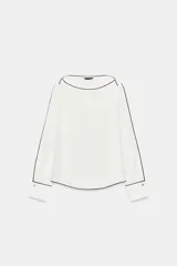 Blusa de crepe con cuello barco y manga larga. Presenta un diseño minimalista en color blanco con detalles de vivos en color negro que recorren el cuello, las mangas y los puños. Los puños incluyen botones decorativos.