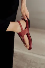 Sandalia plana de cuero color rojo, estilo chinela, con tiras finas adornadas con microtachas doradas. Posee tira de sujeción al tobillo con hebilla.