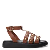 Sandalias estilo gladiador con tiras de cuero color marrón, decoradas con tachas metálicas doradas. Posee una base de plataforma plana color negro y cierre con hebilla ajustable en el tobillo.