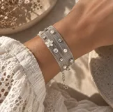 Pulsera multi-hilo color beige con abalorios de perlas, cuentas metálicas plateadas, dijes de cristal y un dije en forma de flor blanca con centro brillante. Cuenta con cadena de extensión metálica.