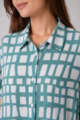 Vestido camisero de silueta relajada, con cuello clásico y cierre frontal de botones. Presenta un estampado geométrico de rectángulos blancos sobre fondo color turquesa.