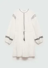 Vestido corto de algodón color blanco con detalles bordados en negro en cuello, hombros y mangas. Presenta mangas largas abullonadas con puños elásticos, cintura elástica con lazo ajustable y diseño evasé.