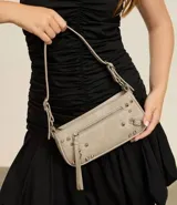 Cartera tipo baguette de PU color beige claro, con acabado liso y detalles de apliques metálicos tipo tachas y argollas en color gunmetal. Presenta un bolsillo frontal con cierre y un tirador de cuero sintético colgante. Incluye asa corta ajustable con hebilla.