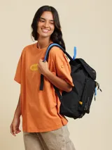 Mochila marrón con detalles en violeta. Tiene un bolsillo frontal superior con cierre, un bolsillo lateral de red y el logo de Rusty Worldwide estampado en naranja.