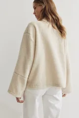 Campera oversized de sherpa suave color beige, con cuello elevado, cierre frontal y bolsillos discretos en las costuras laterales. Presenta hombros caídos y mangas largas amplias.