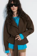 Cazadora trench corta color marrón, confeccionada en mezcla de algodón, con cuello solapa, cierre cruzado con botones, bolsillos de vivo en el delantero y cinturón ajustable del mismo tejido.