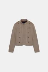 Blazer entallada color beige con rayas verticales finas, cuello redondo y manga larga. Presenta trabillas en los hombros y botones metálicos decorativos en el frente. Cierre frontal con cremallera y forro interior.
