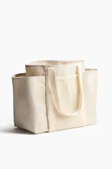 Cartera shopper grande confeccionada en tejido grueso de mezcla de yute color beige, con doble asa de hombro, compartimentos de malla en el frente y dorso, y bolsillos laterales abiertos.