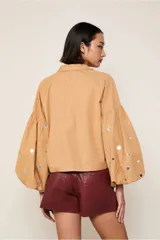 Camisa color beige de corte crop, con cuello camisero, mangas largas abullonadas con detalle de espejos y cierre frontal con botones.