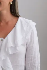Blusa blanca de manga larga con volados en el cuello y en los puños. Presenta un cierre frontal con botones y un diseño fluido.