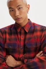 Camisa de franela a cuadros rojos, negros y azules, de corte recto, manga larga, con cuello clásico, cierre frontal con botones, dos bolsillos con solapa abotonada en el pecho y puños abotonados.