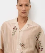 Camisa de lyocell color beige con corte boxy, cuello bowling y mangas cortas. Presenta un delicado bordado floral artesanal y cierre frontal con botones.