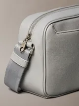 Cartera tipo cámara gris claro de poliuretano texturizado, con cierre superior de cremallera, correa de hombro ajustable y monograma CK grabado en la esquina delantera.