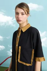 Túnica de algodón color negro, con cuello polo, manga corta, cartera frontal con botones y bolsillo ojal en el pecho. Presenta detalles de voladitos mostaza en el cuello, la botonera, los puños, el bolsillo y el ruedo.