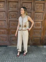 Chaleco beige sin mangas con escote en V y cierre frontal mediante lazos de atar. Presenta bordados florales en color marrón y terminaciones con bordes ondulados en contraste.