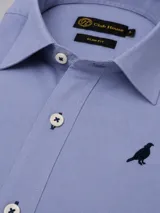 Camisa de algodón color celeste, corte slim fit, con cuello clásico, botones blancos con costura azul y un bordado de un pájaro en el pecho.