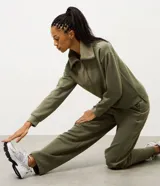 Buzo deportivo de corte oversized en color verde oliva, confeccionado en tejido modal con mezcla de poliéster y elastano. Presenta cuello alto con cierre frontal de cremallera y paneles frontales con textura acanalada. Incluye mangas largas con puños elásticos ajustados.