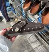 Zueco de gamuza marrón oscuro, forrado en cuero vacuno, con tira ajustable con hebilla metálica decorativa y tachas plateadas alrededor de la suela.