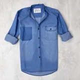Camisa de jean celeste con cuello mao, manga larga con puños enrollados, cierre con botones y dos bolsillos frontales, uno de parche y otro con solapa y botón.