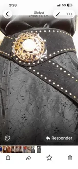 Cinto de cuero agamuzado color negro, con detalle de tachas metálicas a lo largo de la tira y hebilla circular grande con diseño calado y esfera central decorativa.