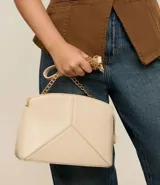 Cartera bandolera pequeña de diseño estructurado en color beige, confeccionada en poliuretano con acabado liso. Presenta un diseño geométrico con costuras en relieve en la parte frontal, cierre superior con cremallera y una elegante correa de cadena metálica en tono dorado.