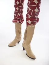 Botas de caña alta de gamuza color tostado, con diseño de estilo western, tacón medio y detalles de pespuntes y flecos decorativos.