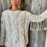 Sweater de lana color crudo con trenzas y tiras de cuero entretejidas. Tiene mangas largas con flecos de cuero en los puños.