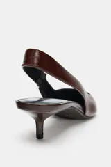 Zapato de tacón tipo destalonado confeccionado en piel con acabado brillante. Presenta diseño de punta fina, pespuntes decorativos en el empeine y tira trasera con elástico lateral. Incluye tacón kitten fino.