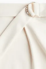 Short de tiro alto color blanco roto, con pinzas frontales y un cinturón fijo con anillas metálicas decorativas en un lateral.